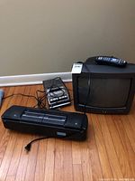 Photo showing all three items: GE dual cassette boom box, Sony cassette recorder, and RCA CRT TV with remote on hardwood floor against beige wall