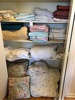Closet shelves with stacks of folded bed linens, pillowcases, blankets, and two decorative cushions at bottom
