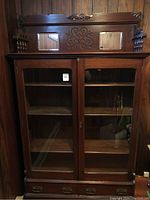 Front view of large wooden display cabinet with glass doors and carved design on top.