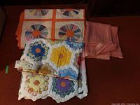 Two handcrafted quilts folded and stacked, with a wool throw blanket to the side, showing colors and patterns of the quilts and blanket material.