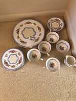 Photo showing full set of dishes including large plates, side plates, teacups, saucers, and creamer jug arranged on carpet.
