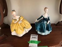 Two Royal Doulton figurines displayed side by side, The Last Waltz in yellow and Janine in teal dress.