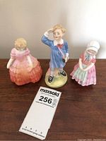 Three Royal Doulton porcelain figurines on a wooden surface, showing full view of each figurine together.