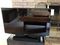 Front view of Samsung 40 inch TV mounted on stand with remote placed on the base stand, screen off reflecting room.