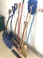 Photo showing the full selection of blue snow shovels, garden shovels, and multiple brooms leaning against a wall.