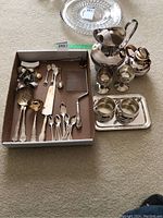 Box with assorted silver plated serving utensils, unmarked water pitcher, small goblets, teapot, sugar bowl, creamer, and small tray.