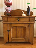 Front view of solid wood washstand with a floral lamp, black vase and white ceramic duck on top. Single drawer with black handle and cabinet door below.