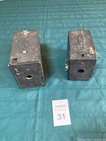 Photo showing two black antique Kodak box cameras side by side on a green surface from the front.