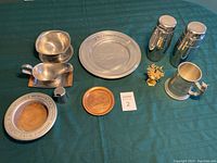 Full overview of lot showing pewter plate, engraved copper plate, two stainless steel cocktail shakers, stainless steel mug with decorative handle, gold-colored sword cocktail skewer set, and other small pewter pieces