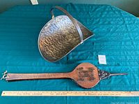 Hammered metal firewood holder with curved handle on blue quilted background, alongside wooden fireplace bellows with stamped tag and ruler.