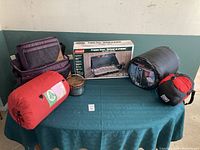 Overview of lot on table: purple/gray cooler bags, Coleman stove box, metal pot and frying pan, gray and red/black compression sacks