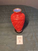 Full view of the carved red lacquer vase from a slight top angle showing the blue glazed interior and gold rim, sitting on a wooden stand.