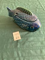 Blue fish-shaped serving bowl with lid and ladle placed on a green cloth, showing side and top views with detailed scales and fins.