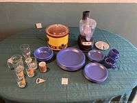 Full view of blue IKEA dishware including plates, bowls, cups, and saucers alongside various drinking glasses, yellow vintage Crockpot, and Black & Decker food processor with accessories.