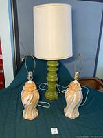 Photo showing two orange and brown ceramic lamp bases without shades and one green ceramic lamp with white cylindrical fabric shade.