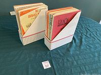 Two stacks of music books in white cardboard holders with grid patterns, arranged on a blue tablecloth.