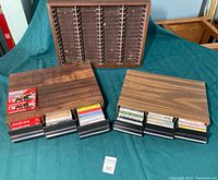 Photo shows two 3-drawer cassette cases laid out with multiple cassette tapes organized in front; a wall-mountable wooden cassette case sits in the background.
