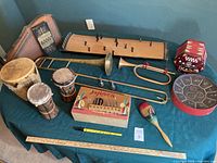 Full view of all musical instruments on a dark teal table, showing trombone, bugle, auto-harp, guzheng, drums, maraca, concertina, and steel tongue drum.