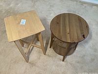 Photo showing both wooden side tables on carpet. Square table is light wood and simple design, round table is medium wood with two shelves.