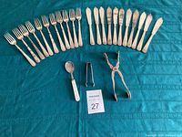 Overview of all items laid out: 10 fish forks, 10 fish knives, 1 child's spoon, 1 pair of tongs on turquoise textile background with MaxSold tag.