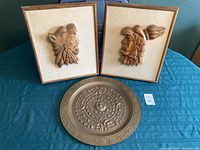 Photo showing two Jose Pinal wood carved warrior head wall hangings and one copper Mayan calendar plate placed on a blue surface.