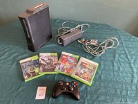 Xbox 360 console, power adapter, A/V cable, wireless controller, and four game cases laid out on fabric surface.