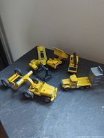Six yellow diecast toy construction and farm vehicles arranged on a dark surface, showing variety and condition.