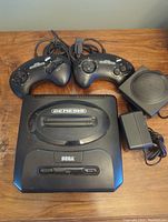 Top view of Sega Genesis console, two wired controllers, AC power adapter, and Menacer adapter on wooden surface.