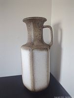Front view of the large vintage pottery vase with gradient brown and cream glaze and handle, standing tall against a white wall.