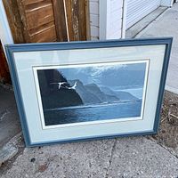 Framed coastal seascape print showing cliffs, ocean, sky, and white-tailed tropic birds in flight. The print is inside a blue painted wooden frame, standing on concrete.