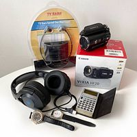 Full lot overview showing all items: Canon VIXIA HF20 camcorder with box, two headphones (TV Ears wireless in packaging and black wired gaming-style), two wristwatches, vintage Casio calculator with case.