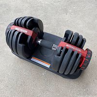 Angled view of Novalook adjustable dumbbell on black plastic base, showing multiple weight plates on each side and red weight selector dial with increments up to 52.5lb.
