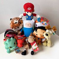 Group of 10 assorted plush toys including a large tiger plush, Mario, Adventure Time Finn, frog, red snake or dragon, gorilla, dog, and others, mostly with tags.