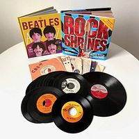 Photo of 17 45 RPM vinyl records including some Beatles singles, a 78 RPM vinyl record, and two hardcover rock-themed books in a white background.