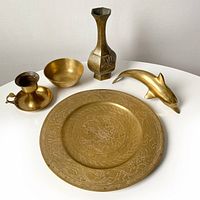 Five brass decor pieces displayed on a white surface: round engraved plate, vase, bowl, dolphin figurine, and candlestick holder