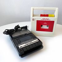 White Radio Shack 'Rugged Recorder' cassette tape recorder with red cassette window and colored control buttons next to black Panasonic slim line cassette recorder with power cord.