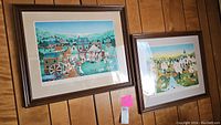 Two framed art prints displayed on a wood-paneled wall showing colorful village scenes with people and horses.