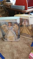 Photo of two boxed Barbie dolls on carpeted floor, one Angel of Peace Barbie with visible golden wings, other Celebration Barbie in gold attire.