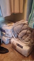 Four blankets and comforters in neutral colors stacked and stored in protective plastic cases, along with one rolled blanket.