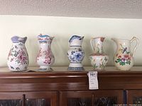 Five Danbury Mint pitchers displayed in a row on top of a wooden cabinet, visible floral and butterfly patterns with colorful, detailed designs.