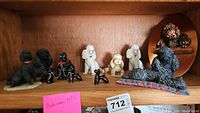 Full display of nine dog figurines on wooden shelf including black, white, and tan poodles and a dog lying on plaid cushion.