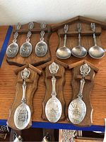 Seven silver-tone decorative German collectible spoons arranged on wooden plaques showing embossed architectural scenes and dates 1980 and 1981.