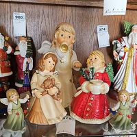 Multiple Goebel Christmas figurines displayed on shelf including angels, children and Santa Claus, arranged with price tags visible.