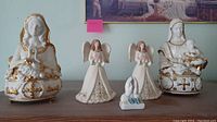 Five religious porcelain figurines displayed on a shelf, including two standing angels, one kneeling angel, a Madonna music box, and Madonna and child statue all with white and gold accents
