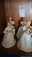 Four ivory porcelain Christmas figurines dressed in elegant gowns with gold accents, arranged on wooden shelf.
