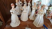 Multiple Lenox Christmas figurines arranged on a wooden shelf, showing the various figures dressed in ivory gowns with gold accents.