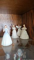 Four Lenox porcelain figurines in evening gowns on a glass shelf in a wooden cabinet.