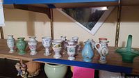 Miniature porcelain vases displayed in a row on a wooden shelf with a green glass basket at the right end.