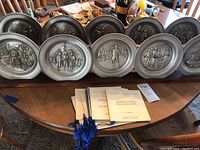 Ten pewter plates displayed on a wooden stand, each featuring raised relief historical scenes from the Bicentennial era. Plates have a matte silver finish and are arranged in two rows, some booklets placed in front of them.