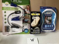 Three small lighting devices in original packaging on table: OttLite LED book light, Nite Ize Nite Note keychain light, and a blue hands-free LED light